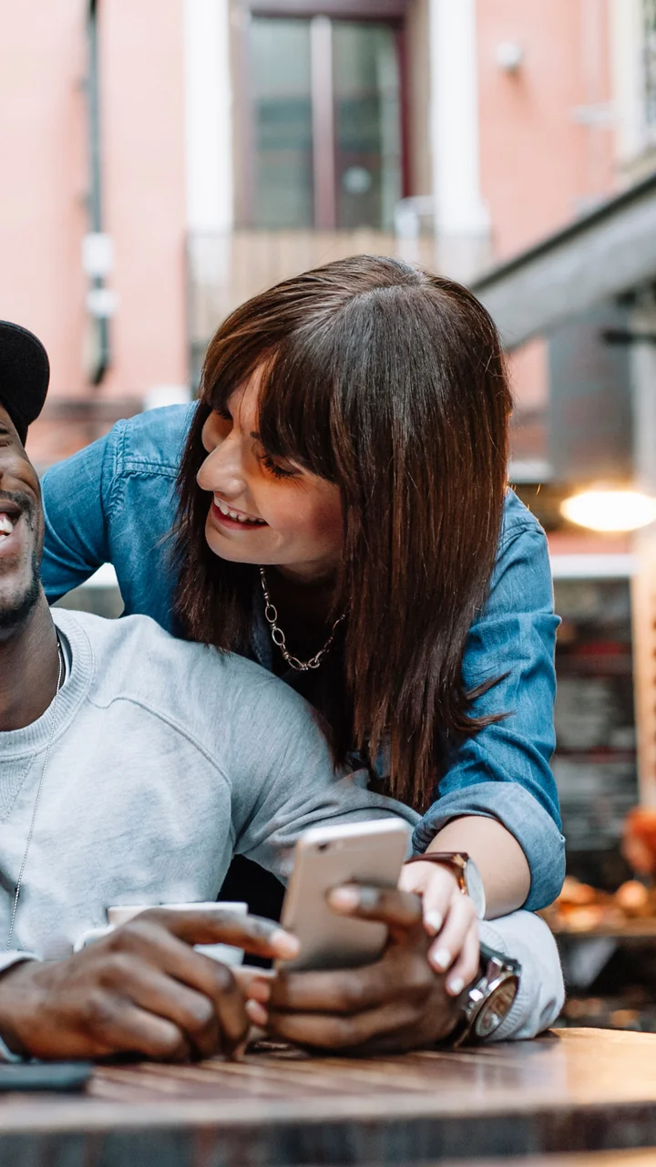 Couple laughing together at an outdoor café table, looking at a phone