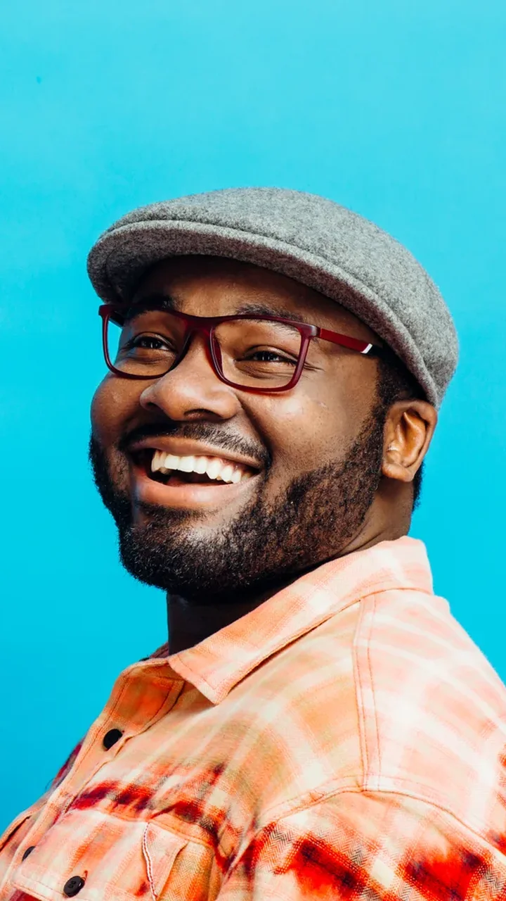 Bearded man in a flat cap and plaid shirt laughing against a solid blue studio backdrop