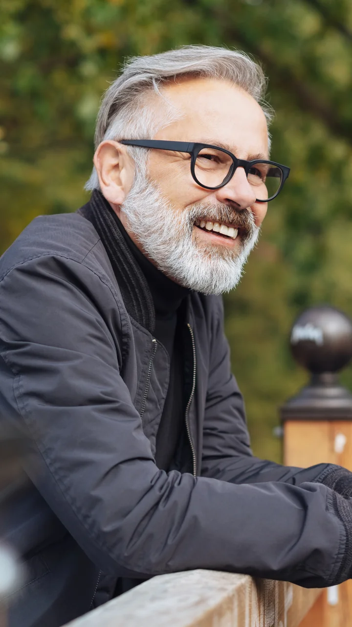Older man with healthy gray hair and glasses laughing while leaning on a park rail