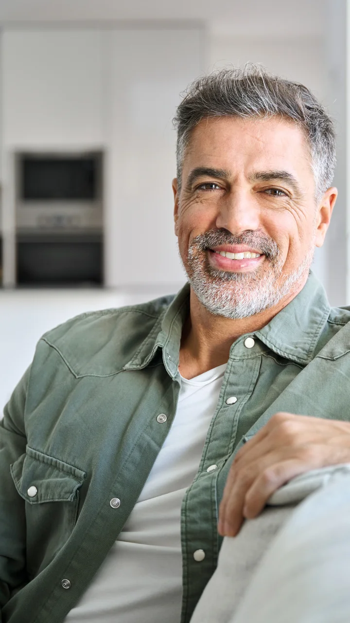 Mature man in an olive shirt seated on a couch smiling at home