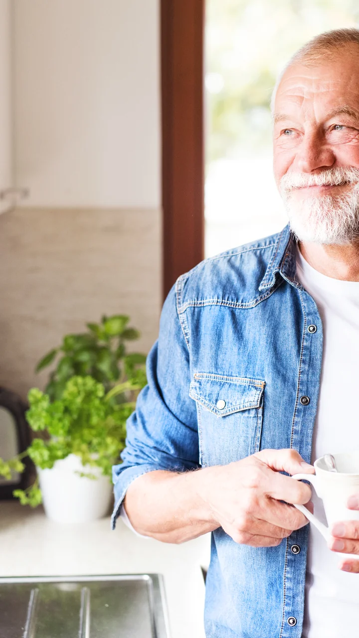 Silver-haired older man with a thick white beard holding a coffee mug in his kitchen